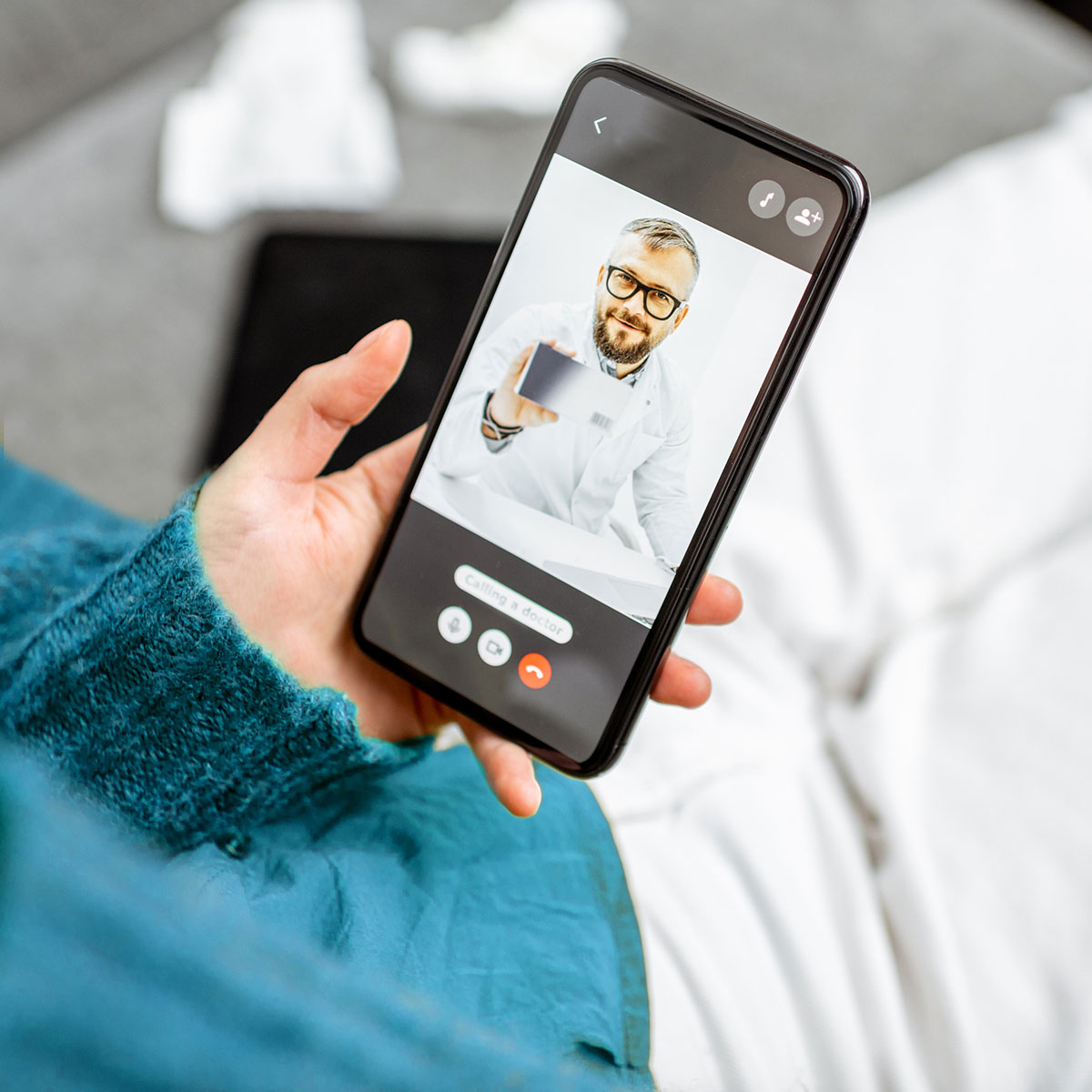 Beratung Stress Hand hält Smartphone mit Videoanruf, auf dem eine Person in weißem Arztkittel ein Tablet zeigt