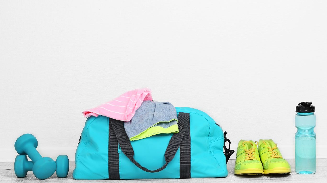 Blaue Sporttasche mit pinkem und grauem Sportshirt, grünen Sportschuhen, zwei blauen Hanteln und einer Wasserflasche vor weißer Wand